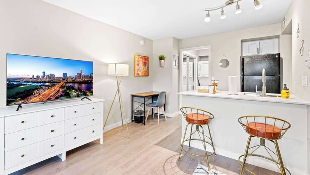 Una cocina moderna con un refrigerador grande, un área de comedor con dos taburetes tipo barra, un televisor montado en un gabinete blanco y un cuadro de un paisaje urbano en la pared.