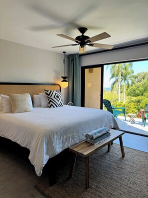 A bedroom with a bed, a ceiling fan, a bench, and a view of greenery outside.