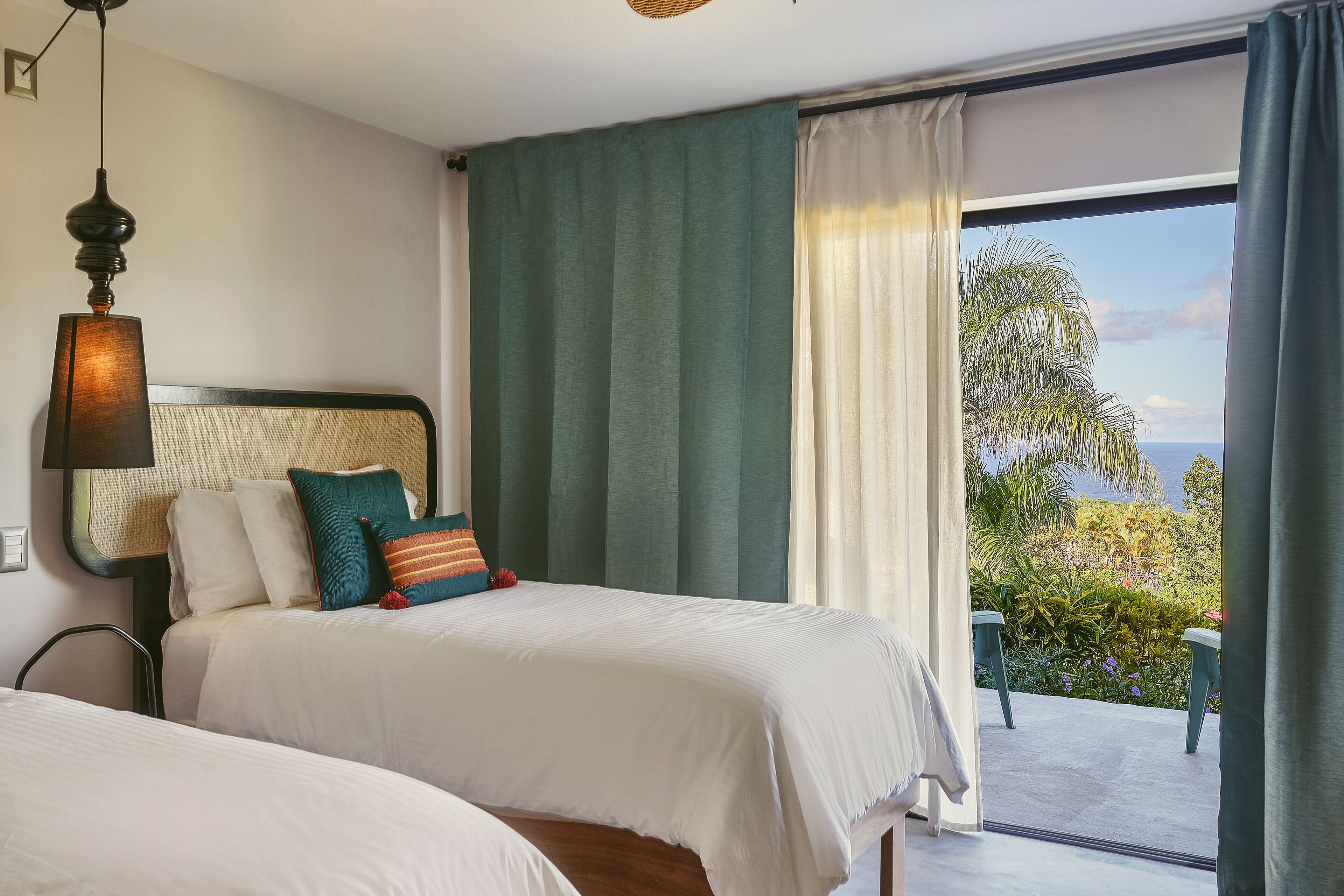 A hotel room with two beds, a large window with curtains, a view of greenery, and a hanging lamp.