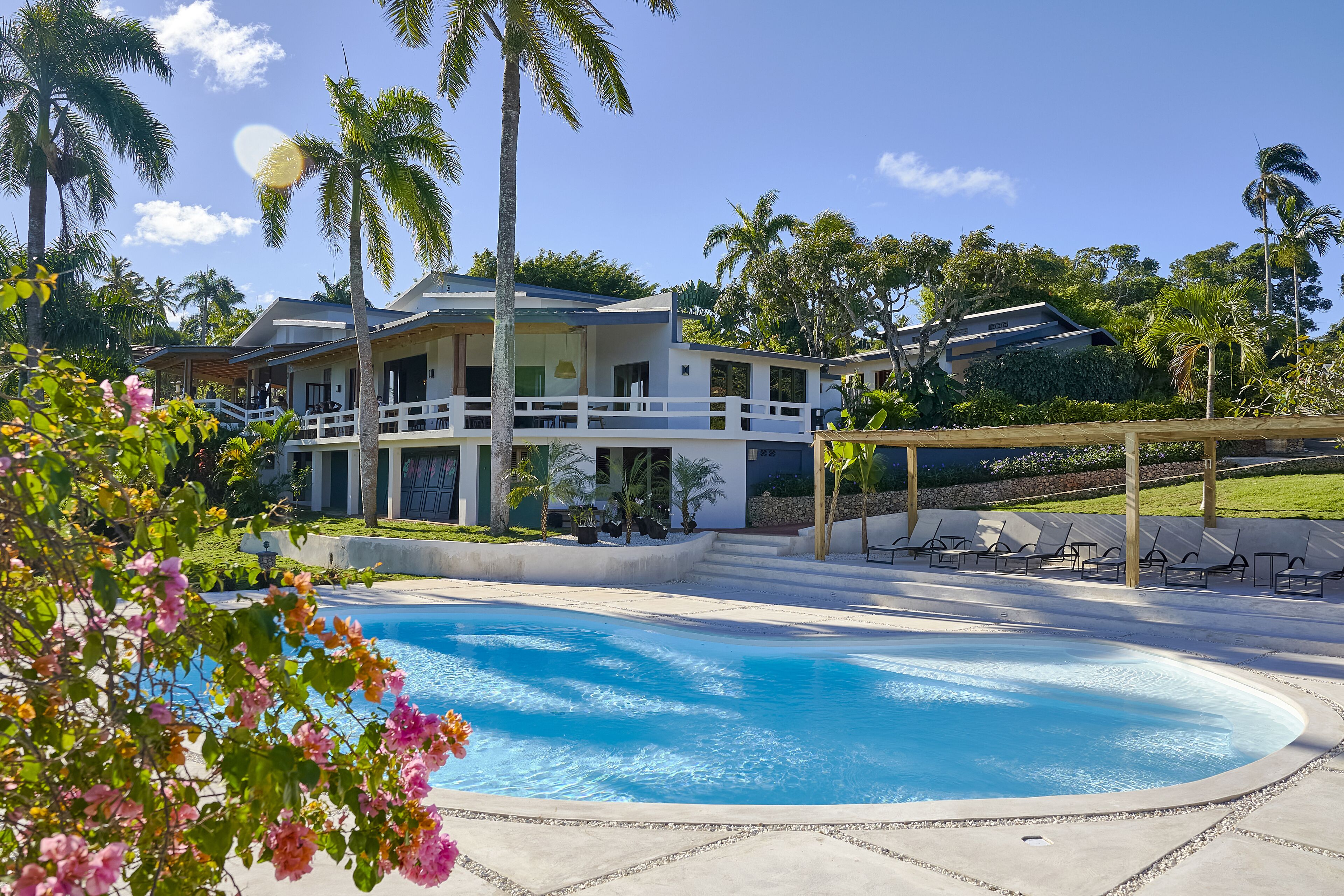 A pool area with a house in the background, surrounded by palm trees and a clear sky.