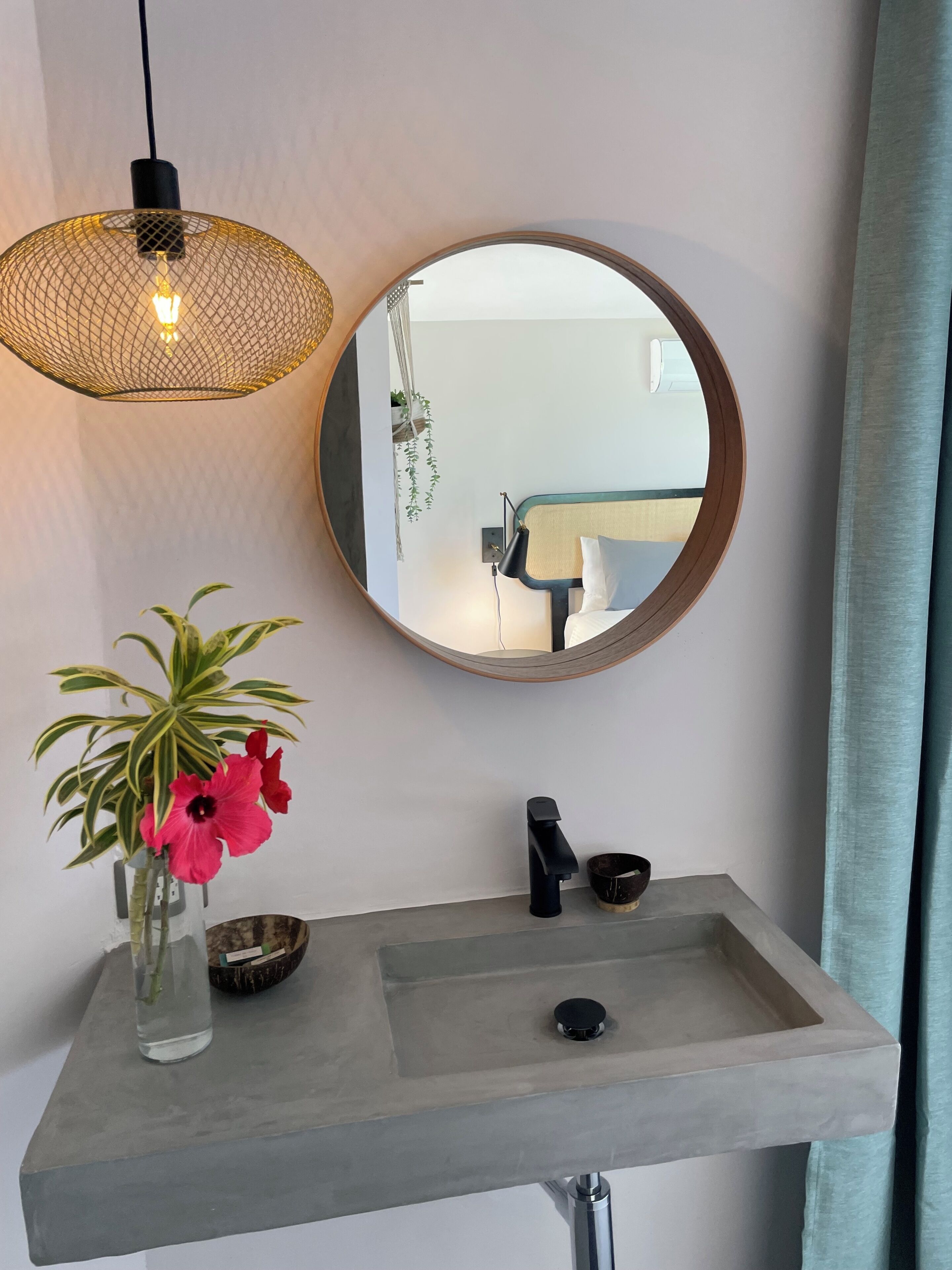 A bathroom with a round mirror, a concrete sink, a hanging light fixture, and a vase with flowers.