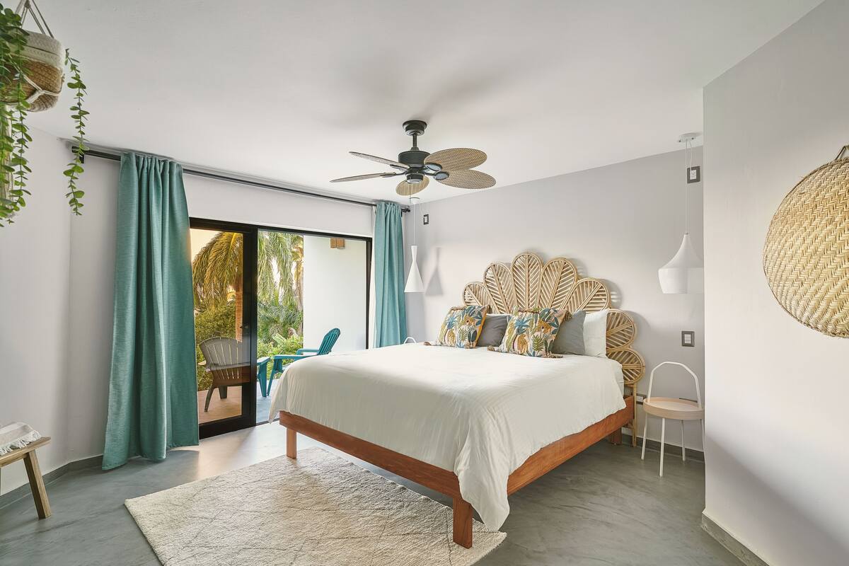 A bedroom with a bed, ceiling fan, and a view to the outside.