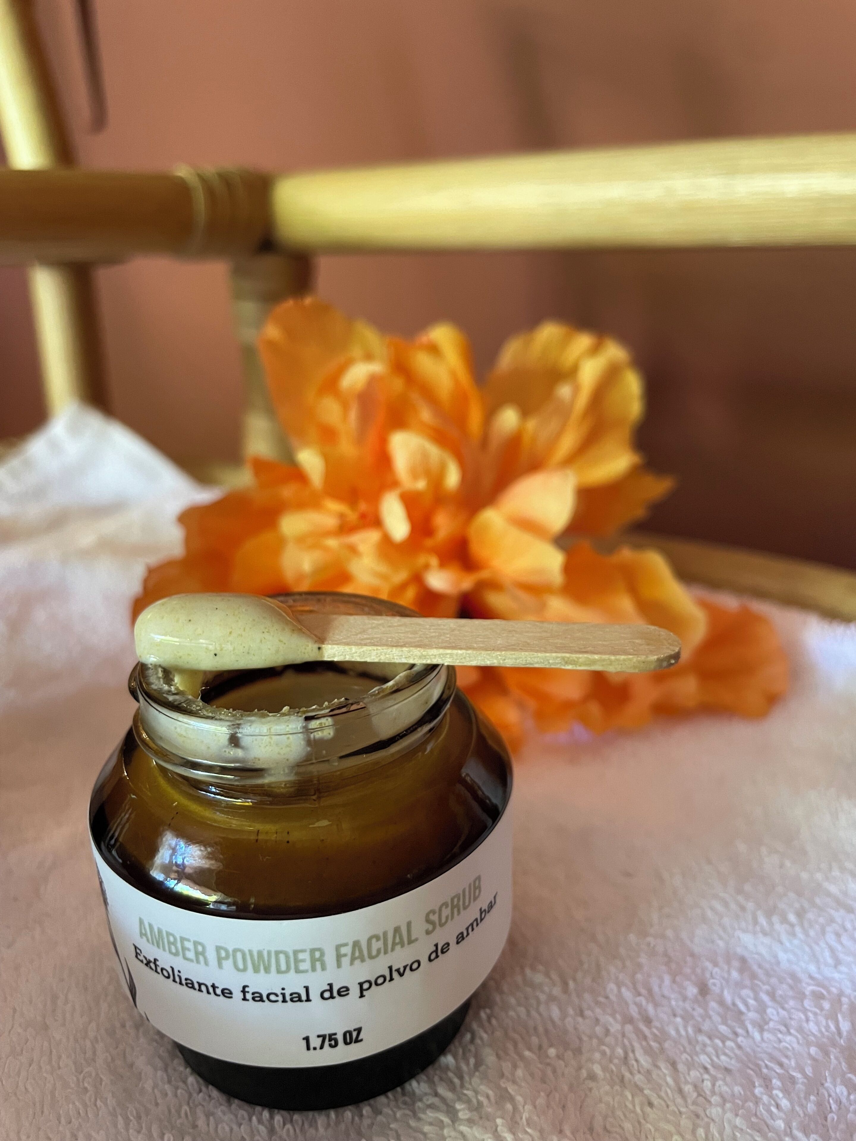Un frasco de exfoliante facial en polvo de color ámbar con una cuchara, sobre una superficie blanca y con una flor de fondo.