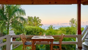 A wooden table and two wicker chairs on a balcony with a view of a lush garden and the ocean at sunset. - Catalina Tropical Lodge (Cabrera)