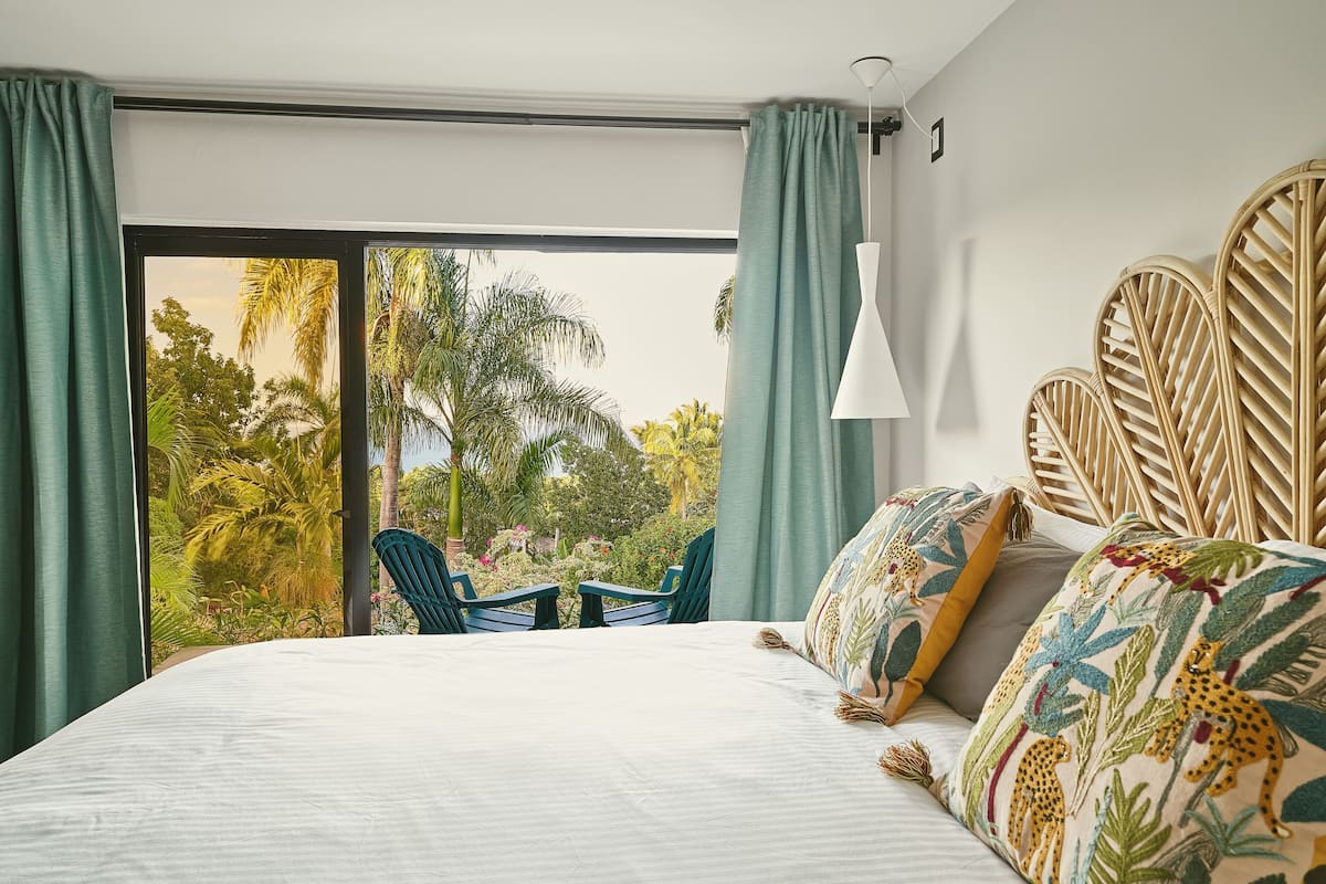 A bedroom with a large bed, two chairs, a view of greenery, and a white pendant light.