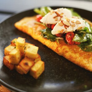 A plate with a slice of omelette topped with tomatoes, greens, and a dollop of cream, accompanied by roasted potatoes.