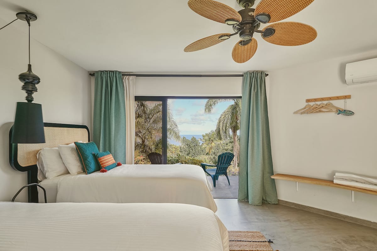A bedroom with two beds, a ceiling fan, a view of the outdoors, and a window with curtains.