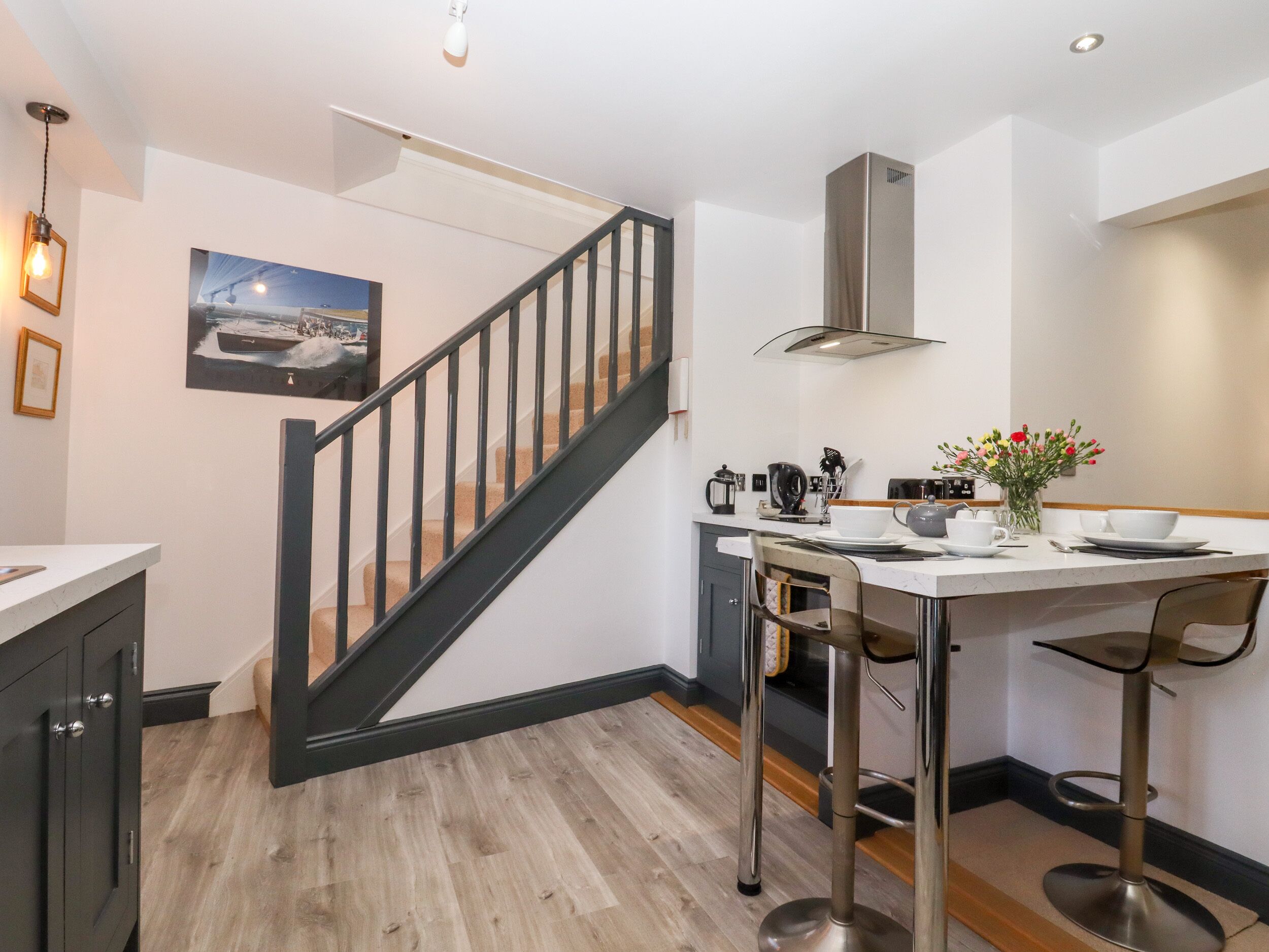 A modern kitchen with a wooden floor, a stainless steel range hood, a dining area with a white table and bar stools, and a staircase with black railings.