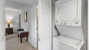 A laundry room with a white washing machine and dryer, a wooden bench, a lamp, and a door leading to another room. - Wailea Elua, #1503 2 Bedroom Condo by RedAwning (Kihei)
