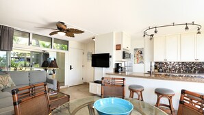 A modern kitchen with a flat-screen TV, a microwave, and a coffee maker. There is a dining area with a glass table and wicker chairs. - Kamaole Nalu, #105 2 Bedroom Condo by RedAwning (Kihei)