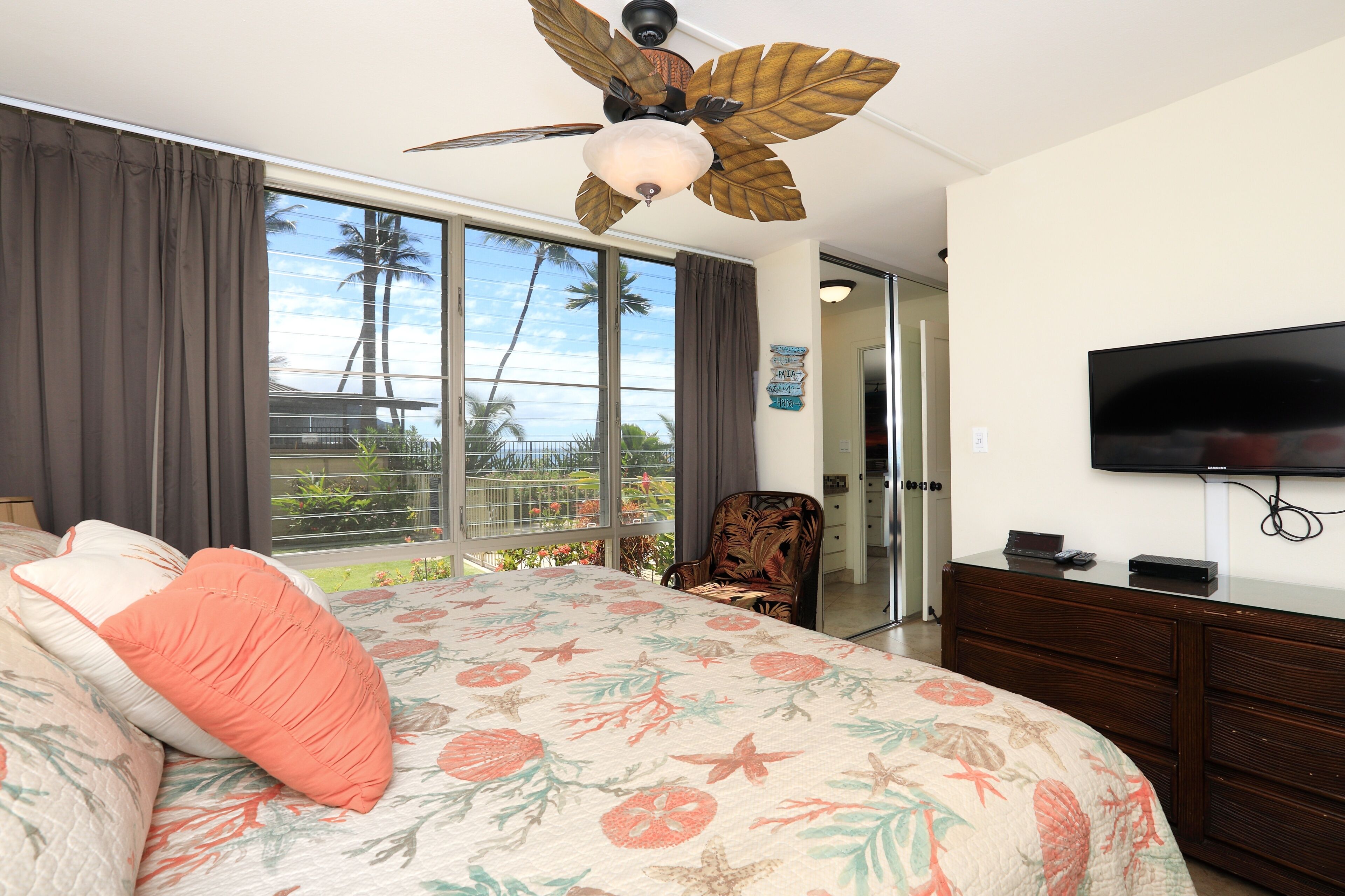 A bedroom with a large bed, a ceiling fan, a television, and a view of palm trees through the window.