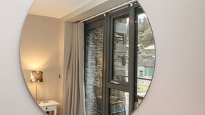 Habitació amb un mirall circular que reflecteix una finestra amb cortines, una petita taula de nit i una làmpada.