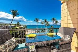 Un balcon agrémenté de meubles en osier, une table dressée avec des assiettes jaunes et des verres à vin, donnant sur une plage bordée de palmiers et surplombant un ciel bleu azur.