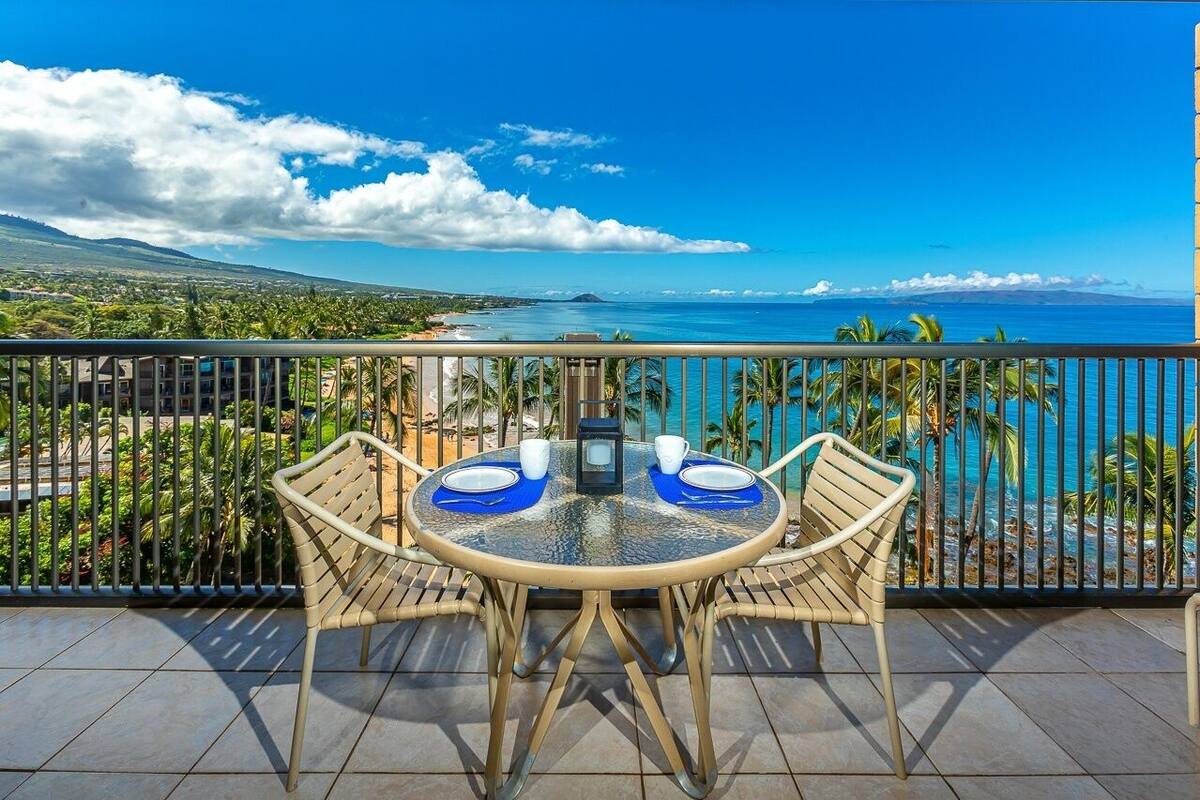 A round table with two chairs on a balcony, overlooking a coastal view with palm trees and a clear sky.