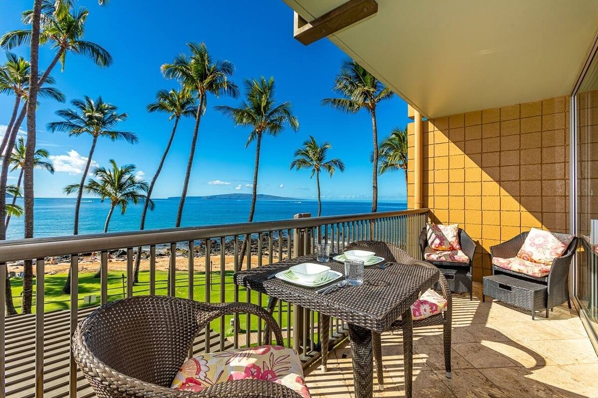 A balcony with wicker furniture, a table with plates and glasses, and a view of the ocean and palm trees.