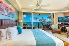 Une chambre d’hôtel avec un lit, un ventilateur de plafond, une télévision et un balcon offrant une vue sur les palmiers et la mer.