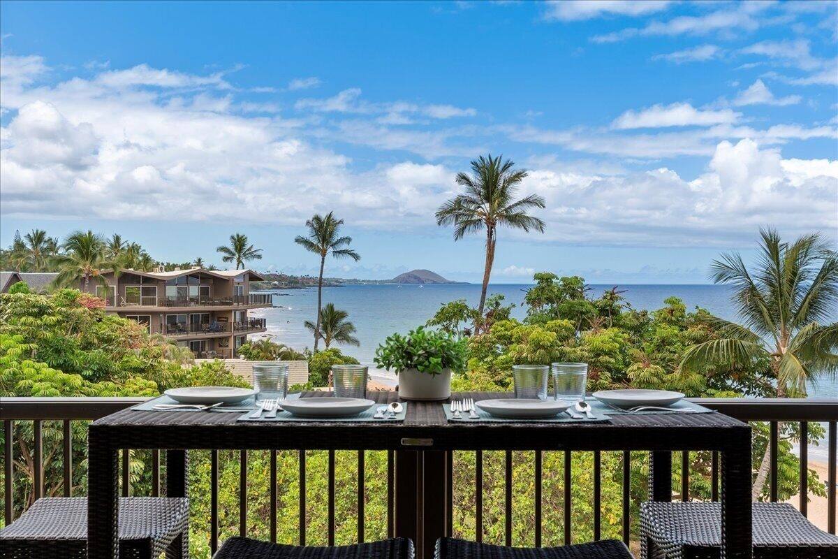 A dining table set on a balcony with a view of the ocean and palm trees.