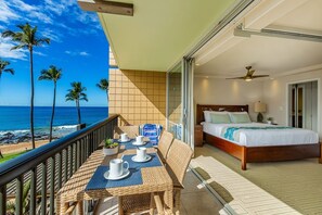 A balcony with a dining table set for two, a bed with a wooden frame, and a view of the ocean with palm trees. - Mana Kai by Coldwell Banker Island Vacations (Kihei)