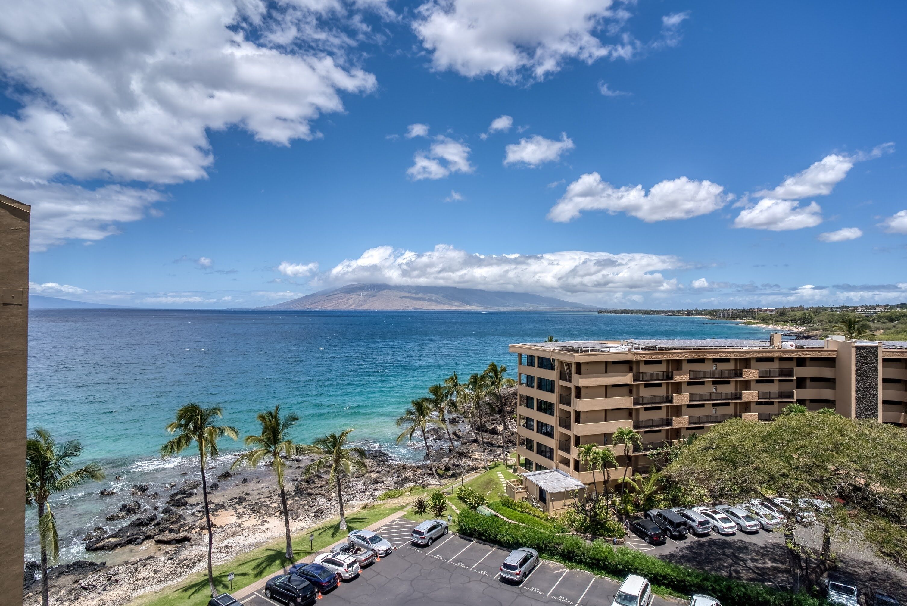 A beachfront hotel with a parking lot, palm trees, and a view of the ocean and a mountain.
