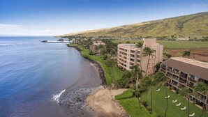 Un hotel costero con varias edificaciones, una playa y aerogeneradores al fondo.