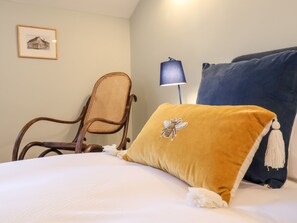 Een bed met een witte dekbedovertrek, een mosterdkleurig kussen met een bijtje, een donkerblauw kussen met een kwast, een rieten stoel en een aan de muur bevestigde lamp.