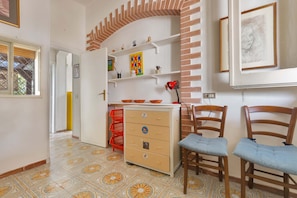 Una habitación con suelo de baldosas, un armario de madera con cajones, dos sillas de madera, una cesta roja y una pared con estantes que contienen varios objetos.