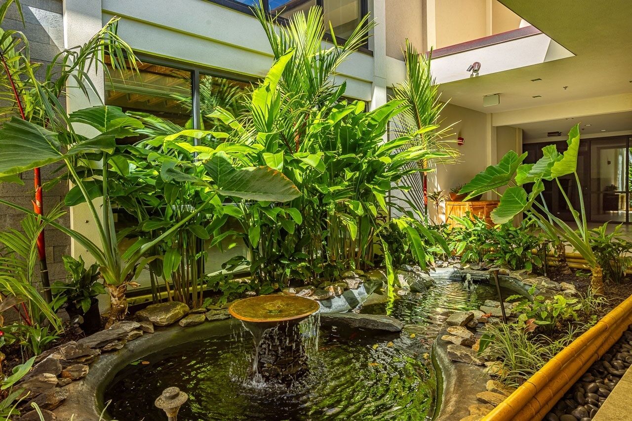 An indoor garden with a pond, lush greenery, and a modern building in the background.