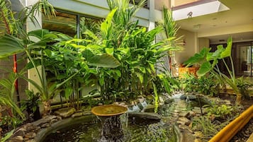 An indoor garden with a pond, lush greenery, and a modern building in the background.