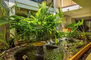 An indoor garden with a pond, lush greenery, and a modern building in the background. - Valley Isle 708 2 Bedroom Condo (Lahaina)