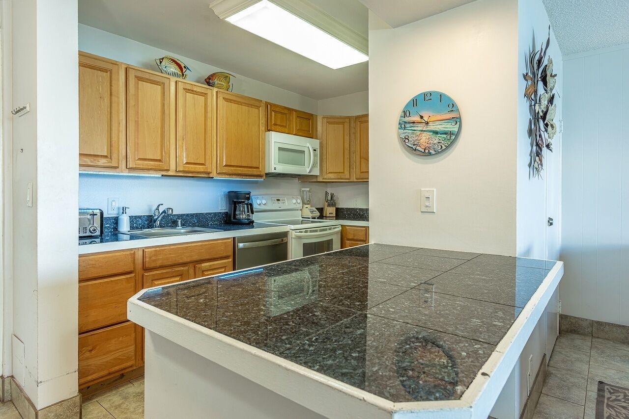 A kitchen with wooden cabinets, a granite countertop, a microwave, a coffee maker, and a clock on the wall.