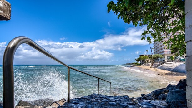 Un escalier menant à une plage où les vagues de l’océan viennent se briser sur le rivage, un ciel bleu dégagé et, au loin, un bâtiment doté de balcons.