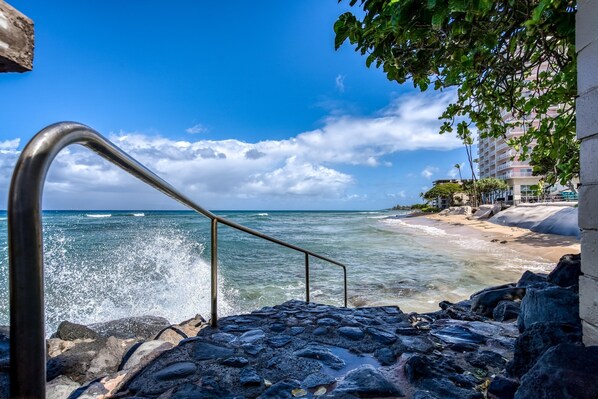 Una escalera que conduce a una playa, con olas rompiendo en la orilla, un cielo azul despejado y un edificio con balcones al fondo.