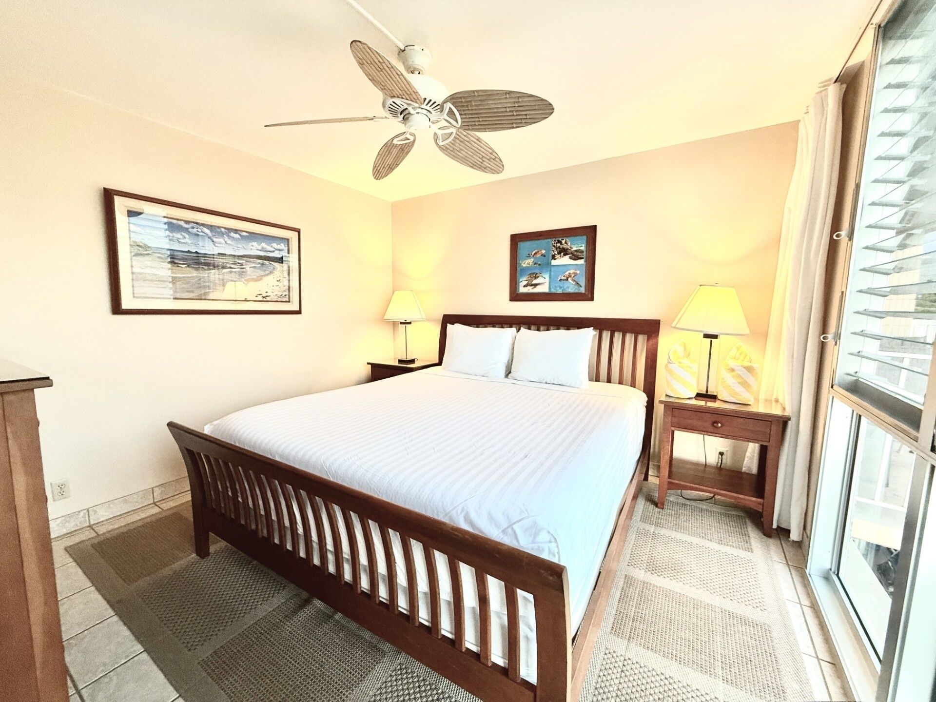 A hotel room with a bed, a ceiling fan, a window with blinds, and two wall-mounted lamps.