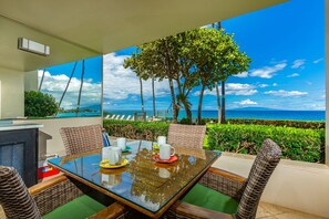 A dining table with chairs, a view of the ocean, and a tree. - Royal Mauian, #114 2 Bedroom Condo by RedAwning (Kihei)