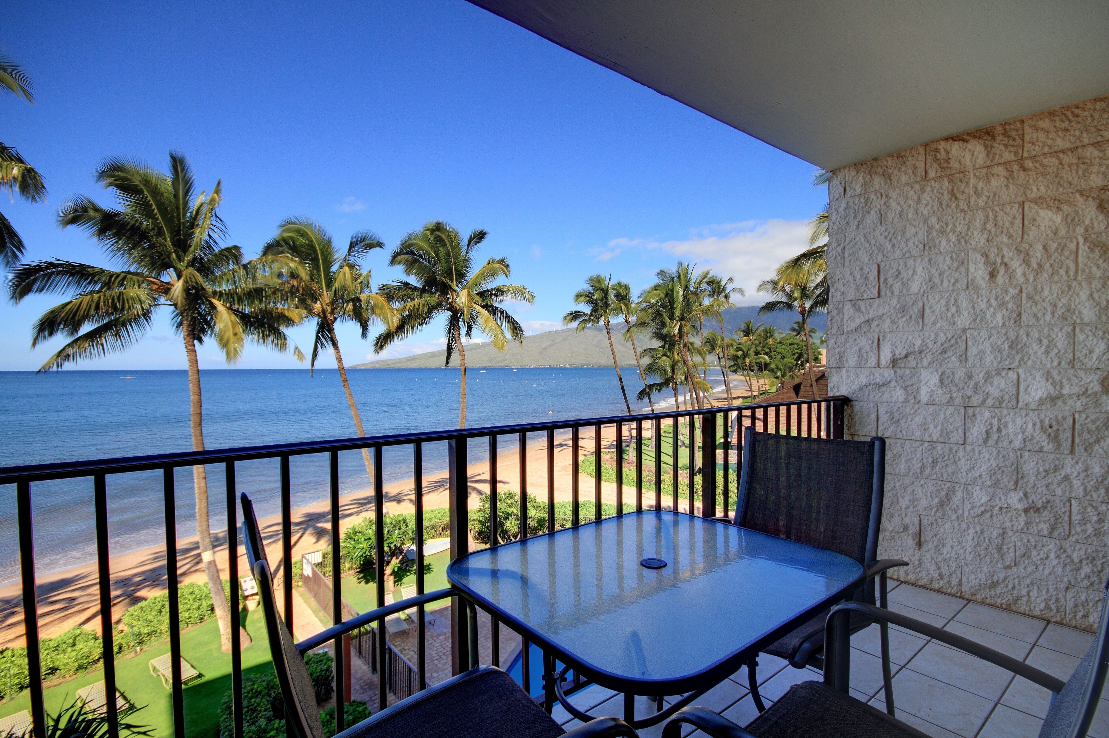 A balcony with a table and chairs, overlooking a beach with palm trees and a view of the ocean.