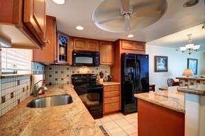 A kitchen with wooden cabinets, a black microwave, a black refrigerator, a stainless steel sink, and a granite countertop. - Kihei Beach, #402 2 Bedroom Condo by RedAwning (Kihei)