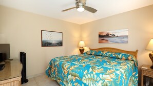 A bedroom with a bed, two bedside lamps, a ceiling fan, and two wall-mounted paintings. - Kamaole Nalu, #102 2 Bedroom Condo by RedAwning (Kihei)