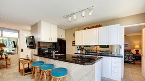 A modern kitchen with a central island, bar stools, and a dining area with a table and chairs. - Kamaole Nalu, #102 2 Bedroom Condo by RedAwning (Kihei)