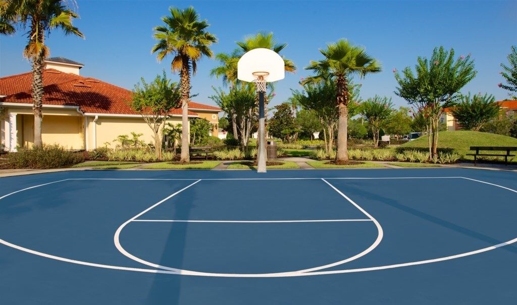 Een basketbalveld buiten, met een ring, omringd door palmbomen en een gebouw met een rood dak.