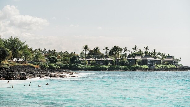 Un hotel frente a la playa con varias edificaciones, palmeras y un litoral rocoso.