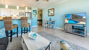 A modern living room with a dining area, kitchen, and a large TV displaying a pool view. - Island Surf by Coldwell Banker Island Vacations (Kihei)