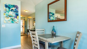 A dining area with a table and chairs, a mirror, and two floral paintings on the wall. - Island Surf by Coldwell Banker Island Vacations (Kihei)
