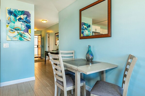 A dining area with a table and chairs, a mirror, and two floral paintings on the wall.