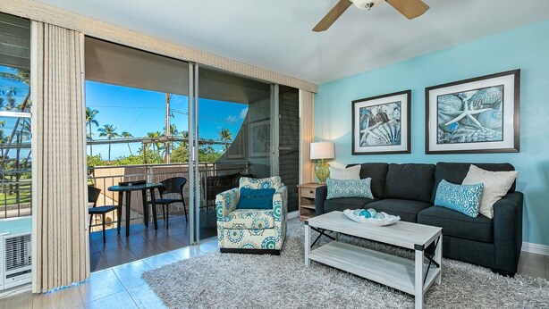 A living room with a sliding glass door leading to a balcony, a dark-colored sofa, a patterned armchair, and a coffee table. - Island Surf by Coldwell Banker Island Vacations (Kihei)