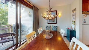 A dining room with a wooden table, white chairs, a chandelier, and a view through a sliding glass door to the outdoors.