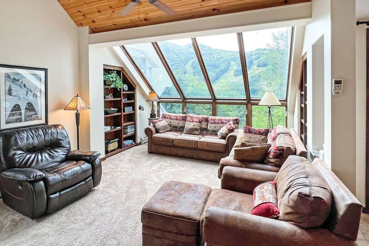 Un salón amplio con techo alto, un ventanal con vistas al exterior, un sofá de cuero, un sillón marrón y una estantería.