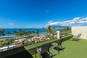 A balcony with artificial grass and two chairs, overlooking a beach and a parking area with several vehicles.
