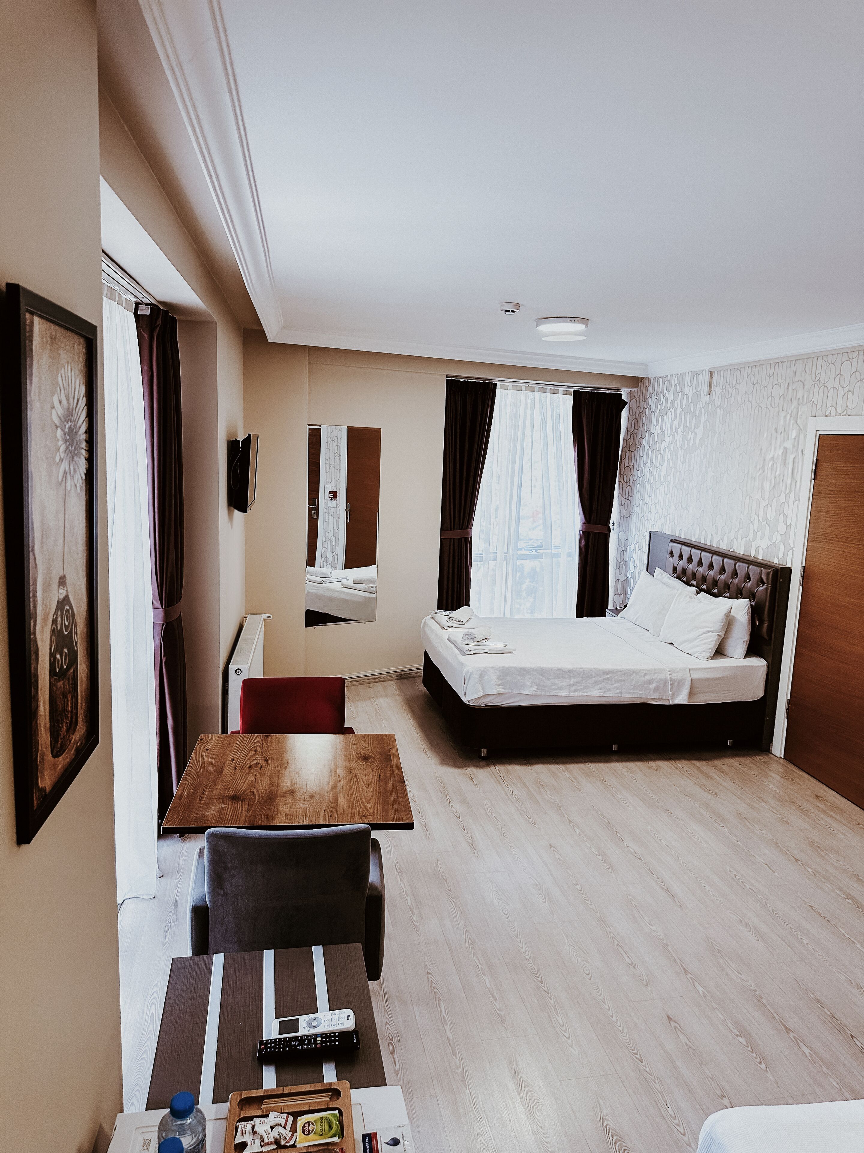 A hotel room with a wooden dining table, a bed with a headboard, a mirror, and a window with curtains.