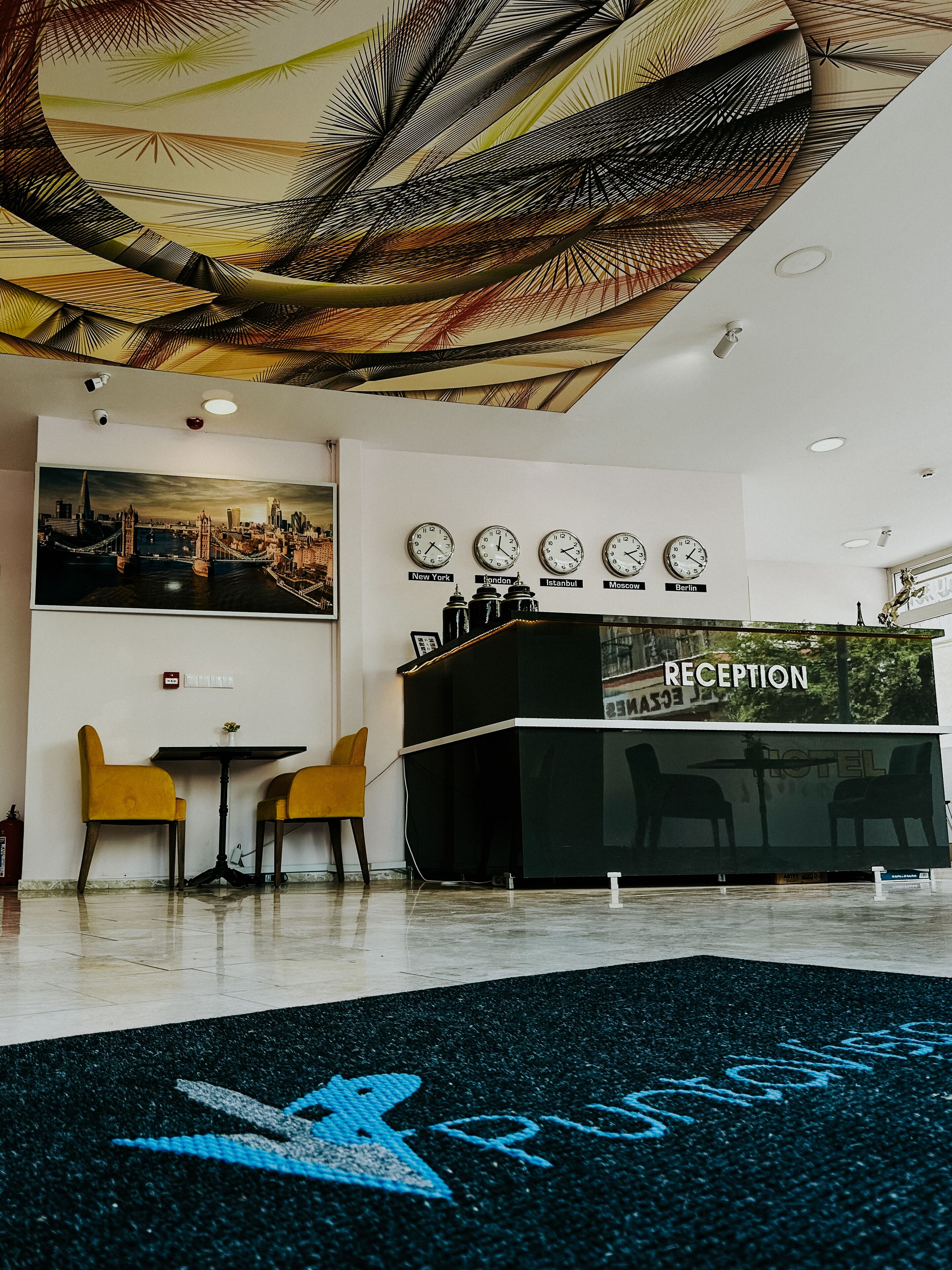 Een hotel receptie met een receptiebalie, twee gele stoelen, een tafel met een vaas en een stadslandschap schilderij aan de muur.