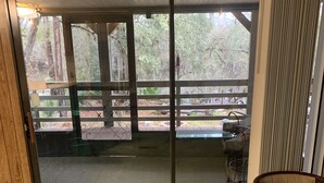Een kamer met een schuifdeur naar een terras met houten balustrade en uitzicht op bomen.